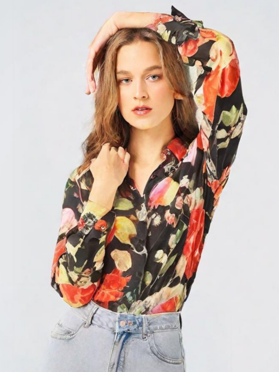 Model wearing oversized black satin shirt with vibrant floral print; light-wash jeans, arms raised, loose wavy hair.