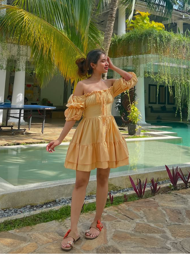 Peach off-shoulder mini dress with puff sleeves and tiered ruffles, by a pool among palm trees; sandals with red flowers.