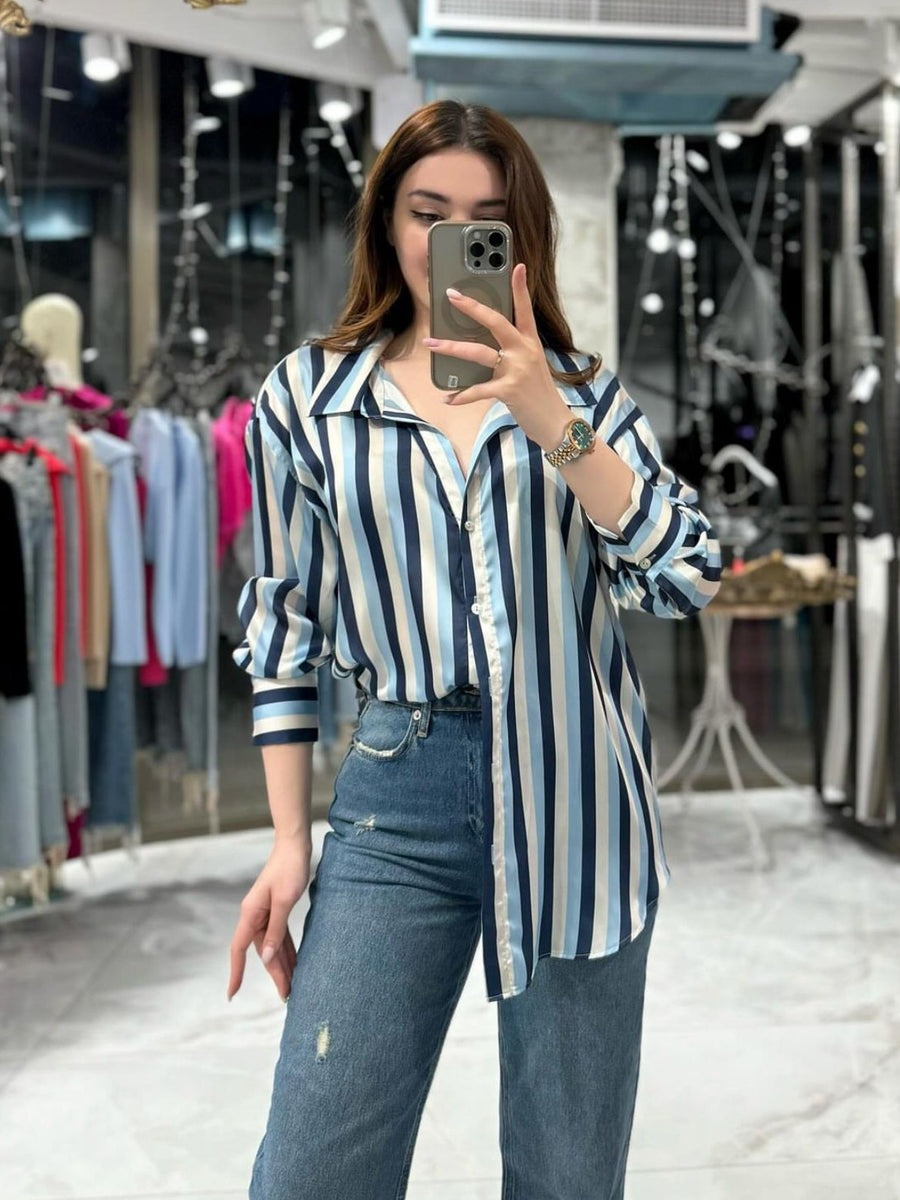 Woman in oversized blue-and-white striped satin shirt with rolled sleeves, jeans, taking a mirror selfie in a boutique.