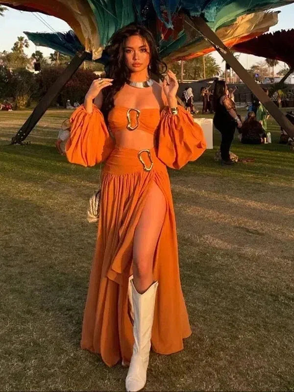 Woman in orange off-shoulder crop top with puff sleeves and matching high-slit skirt with ring buckles, white boots.