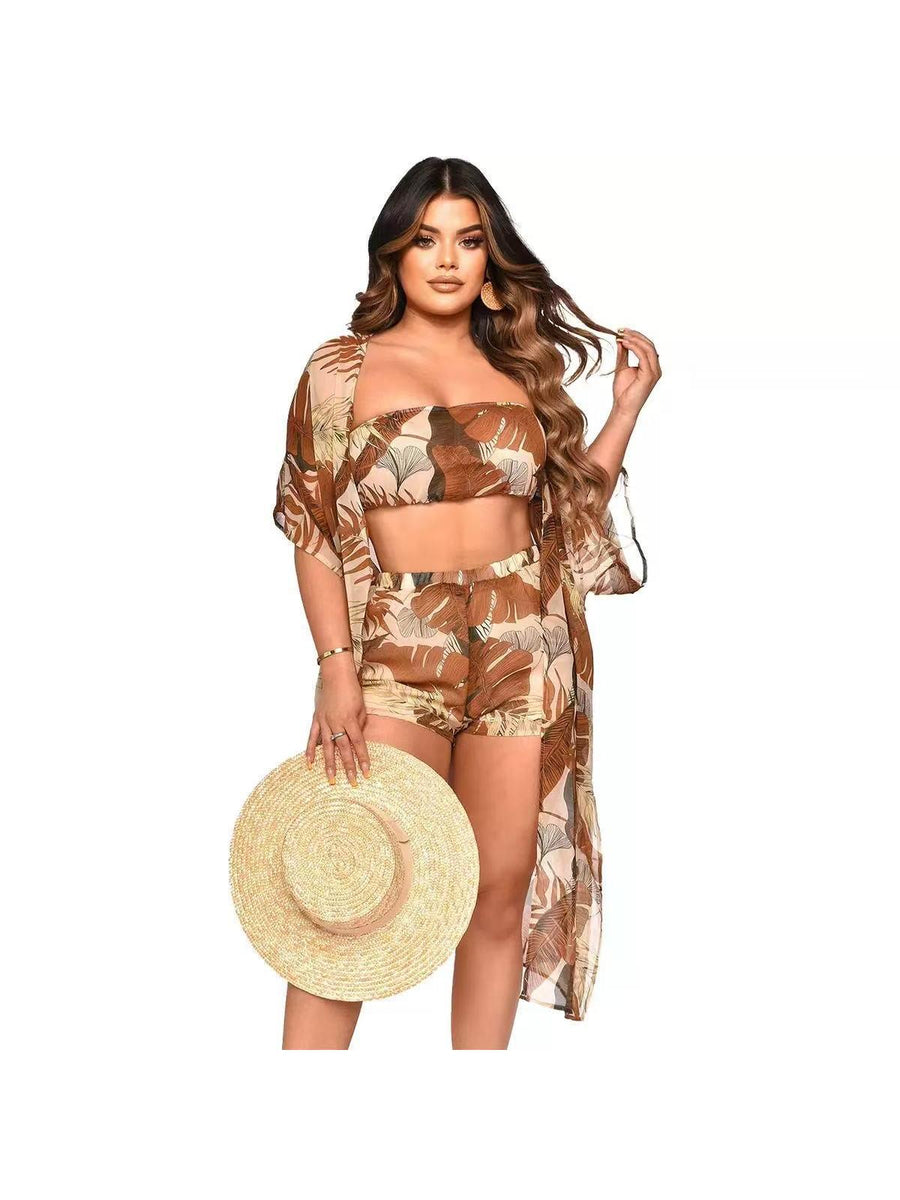 Plus-size model wearing brown tropical leaf print bikini with bandeau top and matching shorts, plus sheer cover-up; holding a wide straw hat.