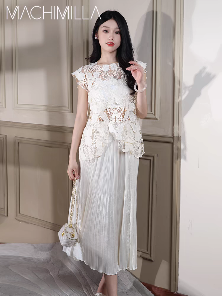 Model in ivory butterfly-cutout lace top over a pleated white midi skirt, holding a small white quilted handbag.