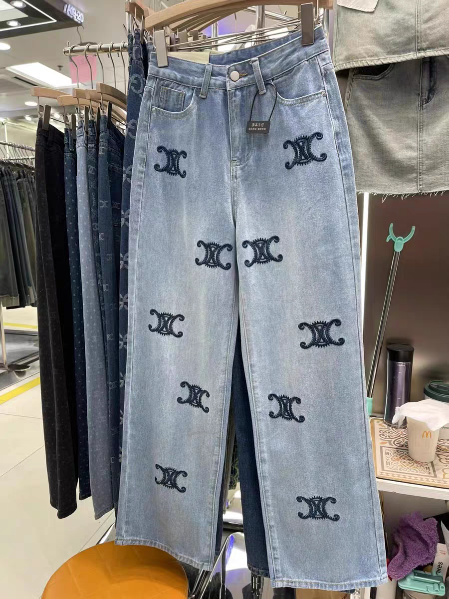 Light-wash distressed wide-leg denim pants with dark blue embroidered logos down the front legs, hanging on a store rack.