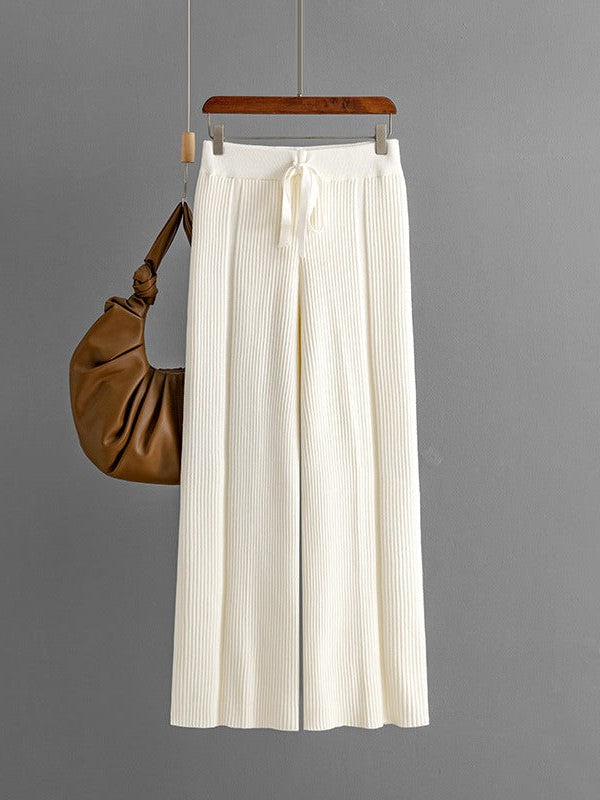 Cream ribbed knit vertical-stripe wide-leg pants with drawstring waist, hanging on a wooden hanger beside a brown bag.