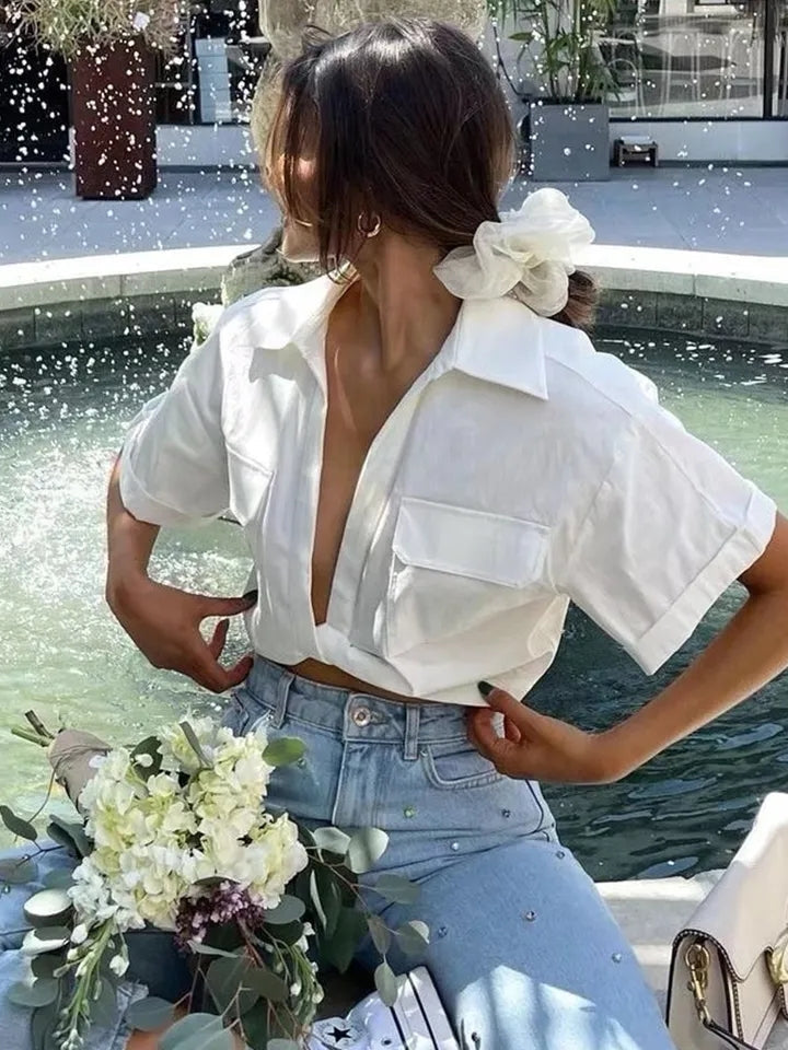 Woman in white linen knotted crop shirt with short sleeves and front pocket, jeans by a fountain, holding a bouquet.