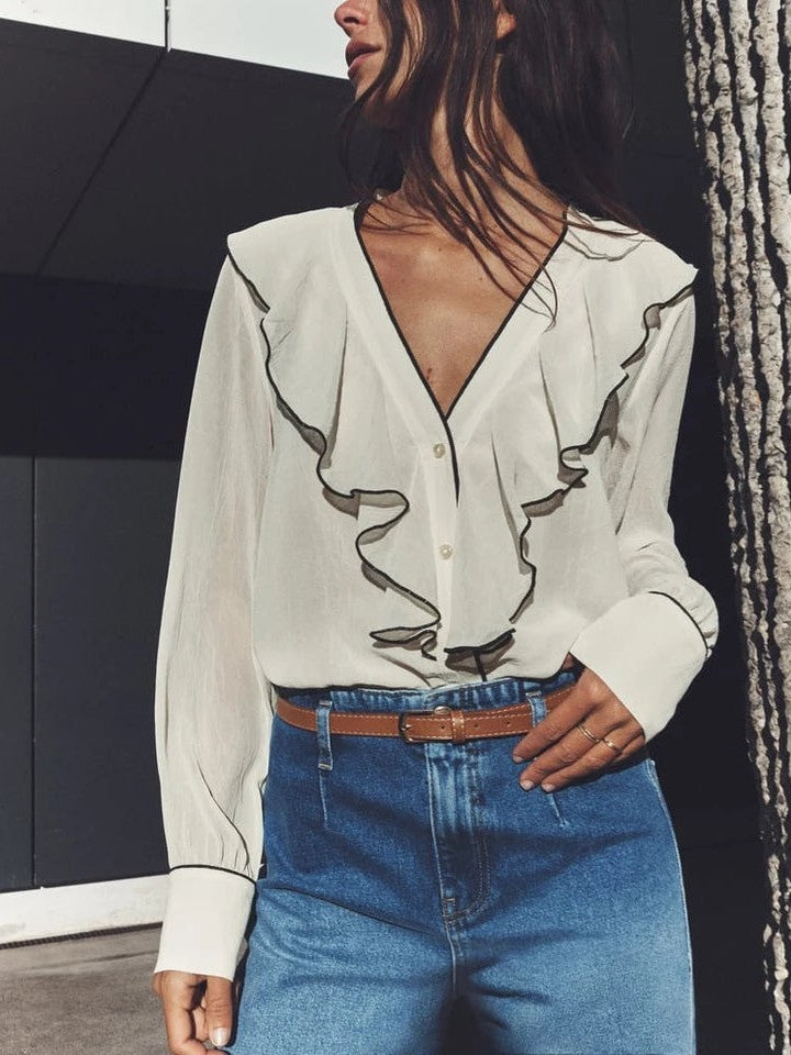 White ruffle-front blouse with black V-neck trim and wide cuffs, gold buttons; tucked into blue high-waisted jeans.