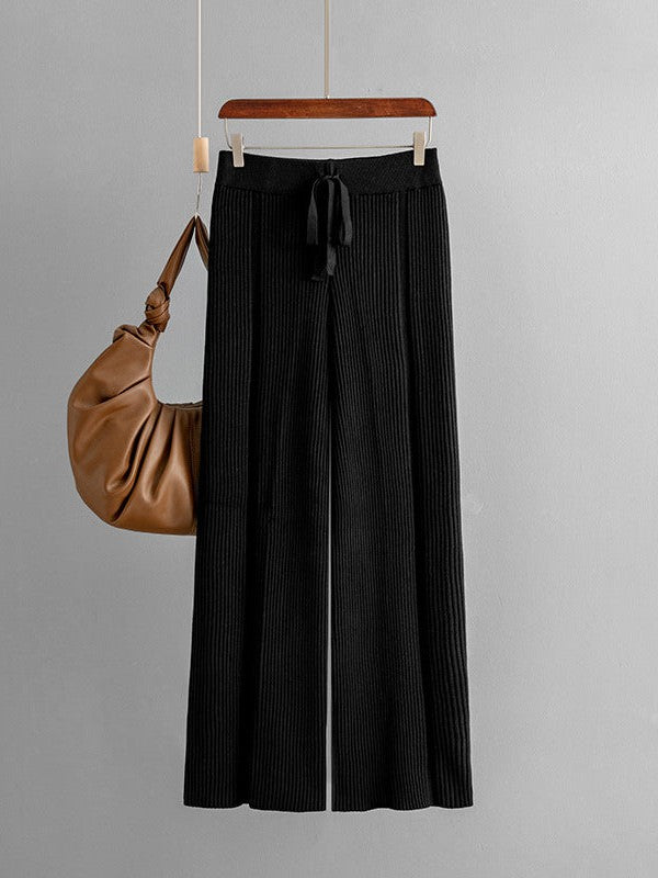 Black knitted wide-leg pants with vertical ribbing and a drawstring waist, hanging on a wooden hanger with a tan bag.