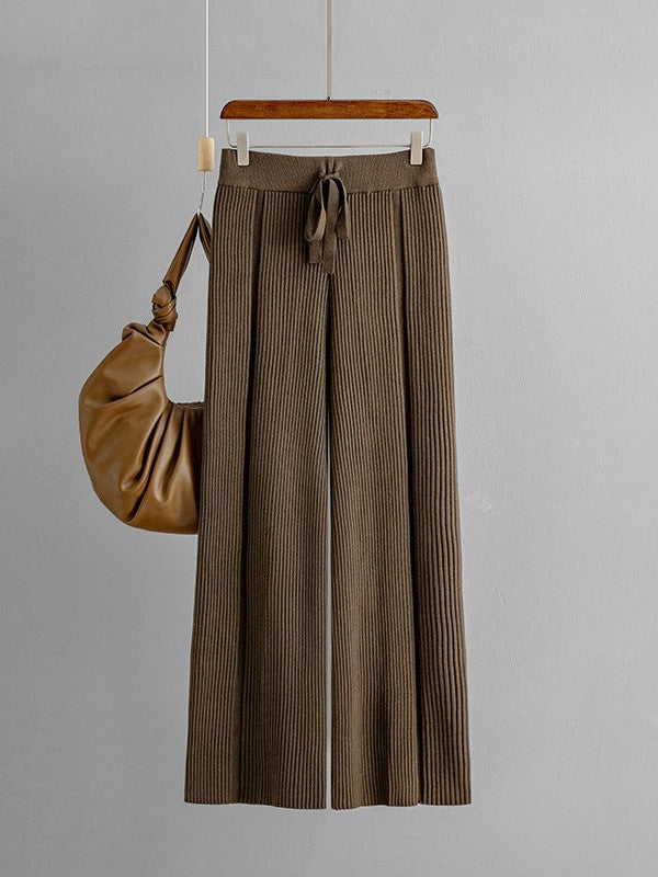 Brown wide-leg knitted pants with vertical ribbing and drawstring waist, hung on a hanger beside a tan leather bag.