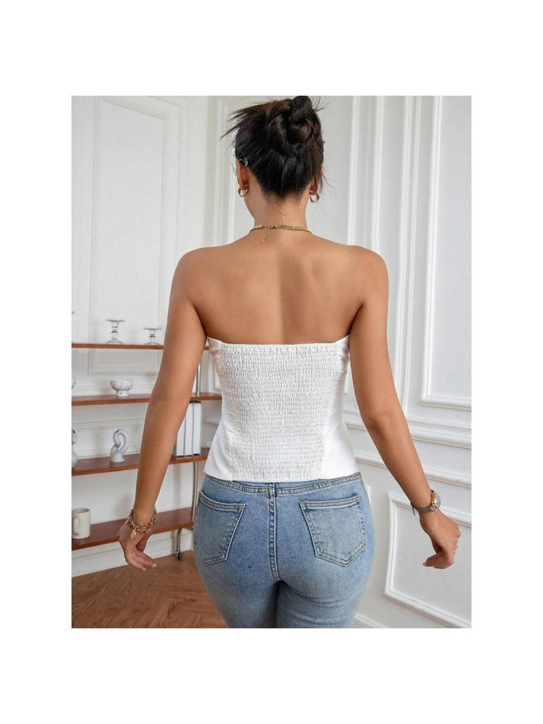 Back view of a woman wearing a white stretchable tube bandeau top with light-wash jeans; hair in a bun, indoors.