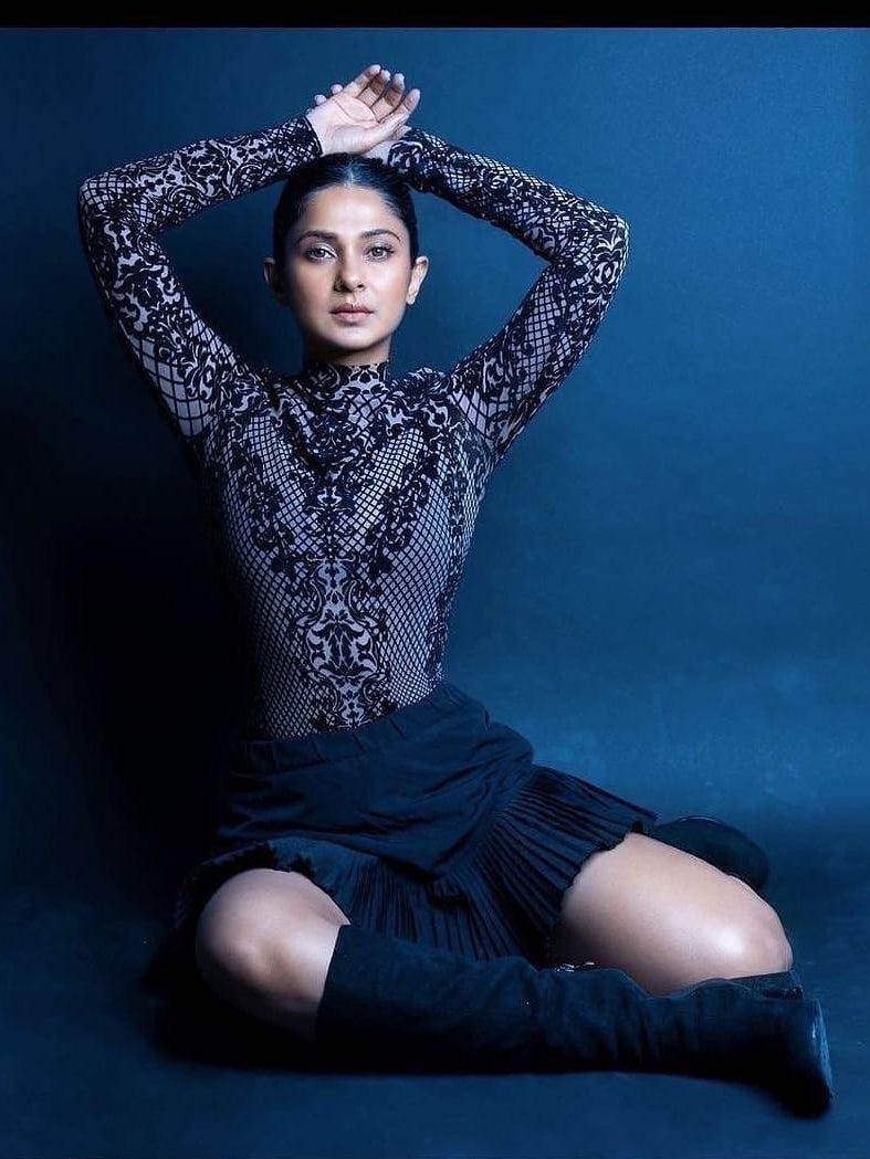 Model in black high-neck mesh velvet lace bodysuit, arms raised, seated against blue backdrop in knee-high boots.