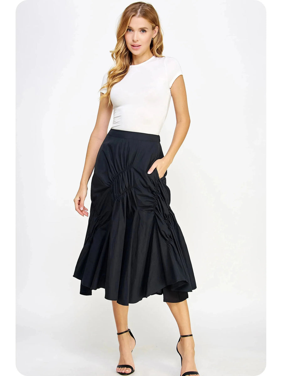 Blonde model in a white fitted top and black A-line pleated ruched ruffle midi skirt with ankle-strap heels.