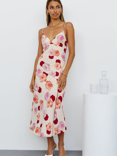 Woman in cream midi dress with pink-red floral print, spaghetti straps, V-neck, side slit, against white wall.