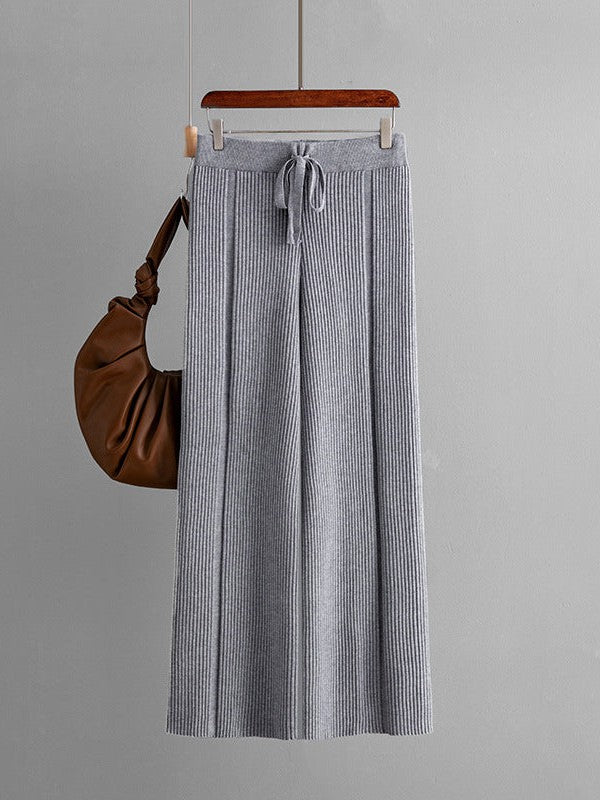 Light gray knitted wide-leg pants with vertical rib stripes, drawstring waist, hanging on a hanger beside a brown bag.
