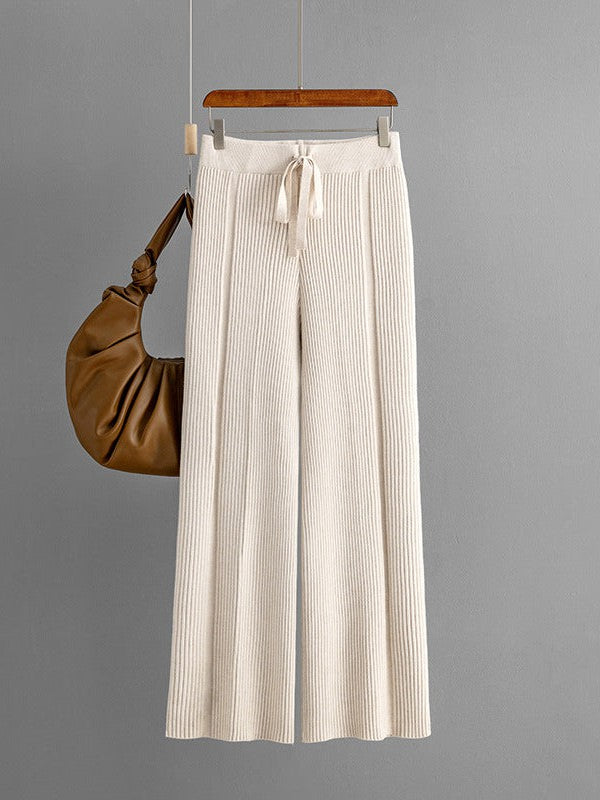 Cream knitted wide-leg pants with vertical ribbing and a drawstring waist, hung on a wooden hanger beside a brown bag.