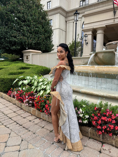 Woman in gray paisley off-shoulder ruffle high-low maxi dress with gold trim, standing beside a fountain.