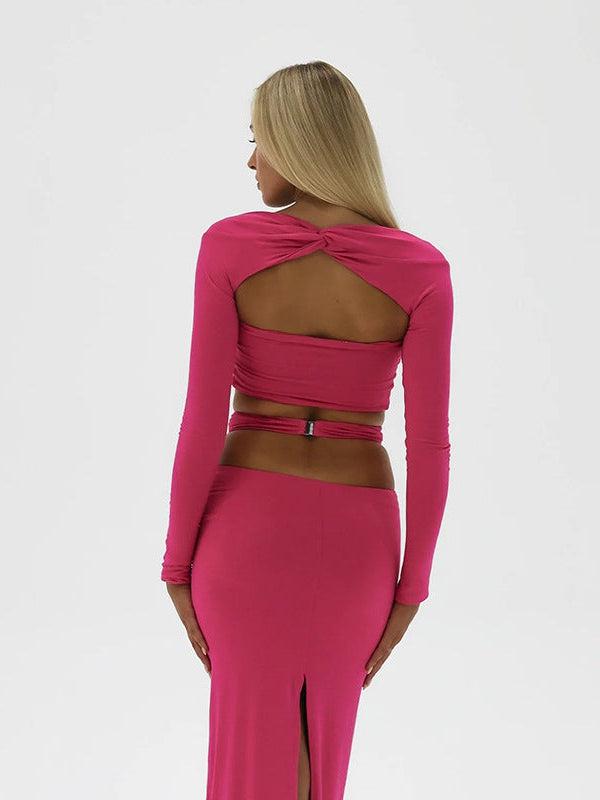 Back view of a model in hot pink long-sleeve cropped top with twisted back cutout and matching skirt with back slit.