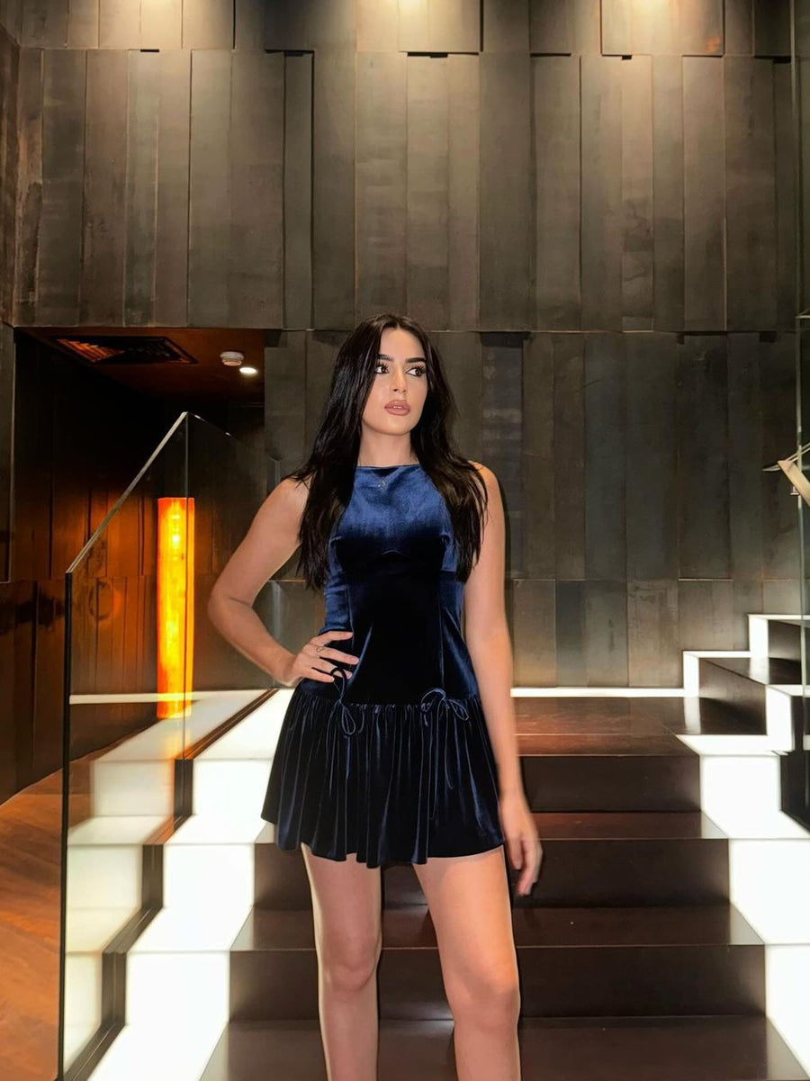Model wearing navy velvet sleeveless mini dress with a bow applique at the waist and a pleated skirt, in a dark lobby.