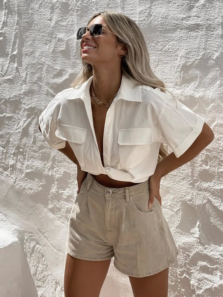 Model wearing white linen knotted crop shirt with rolled sleeves and two chest pockets, beige shorts, sunglasses.