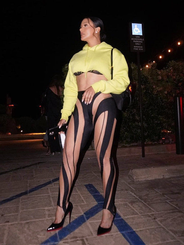 Nighttime sidewalk scene: woman in neon yellow cropped hoodie and black high-waist leggings with sheer panels, red heels.