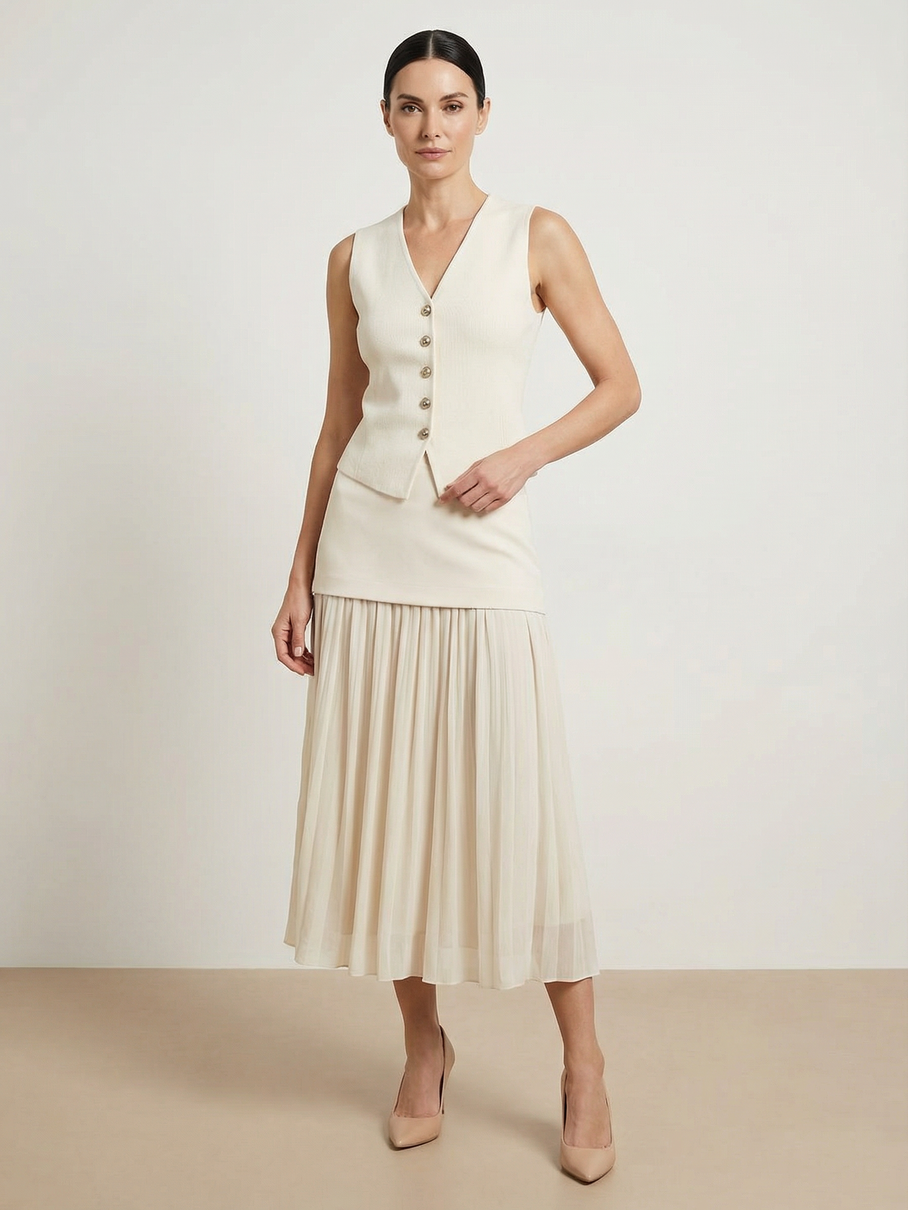 Cream knitted sleeveless vest with V-neck and row of buttons over ivory pleated high-waist midi skirt; nude heels.