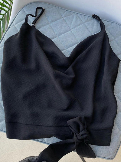 Black spaghetti-strap cowl-neck knot crop top with a draped front and side tie at the hem, laid on a blue quilted surface.