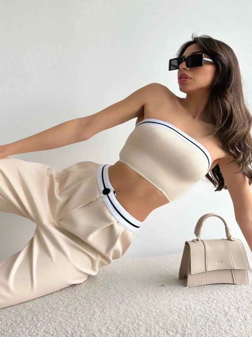 Beige tube top and wide-leg pants with striped waistband, woman in black sunglasses lounging on a cream bed beside bag.