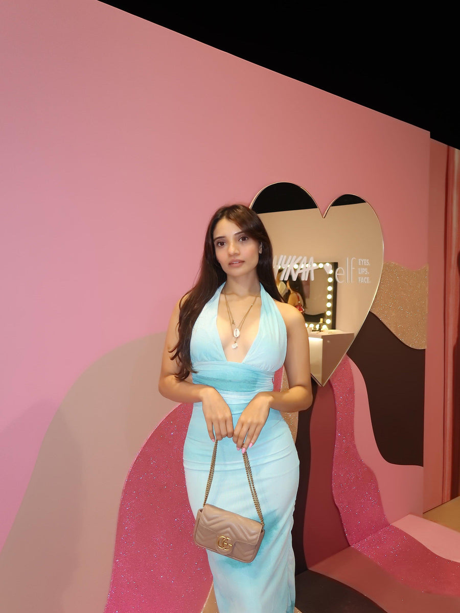 Woman in light blue halter-neck dress with plunging neckline, carrying beige quilted chain bag against pink backdrop.