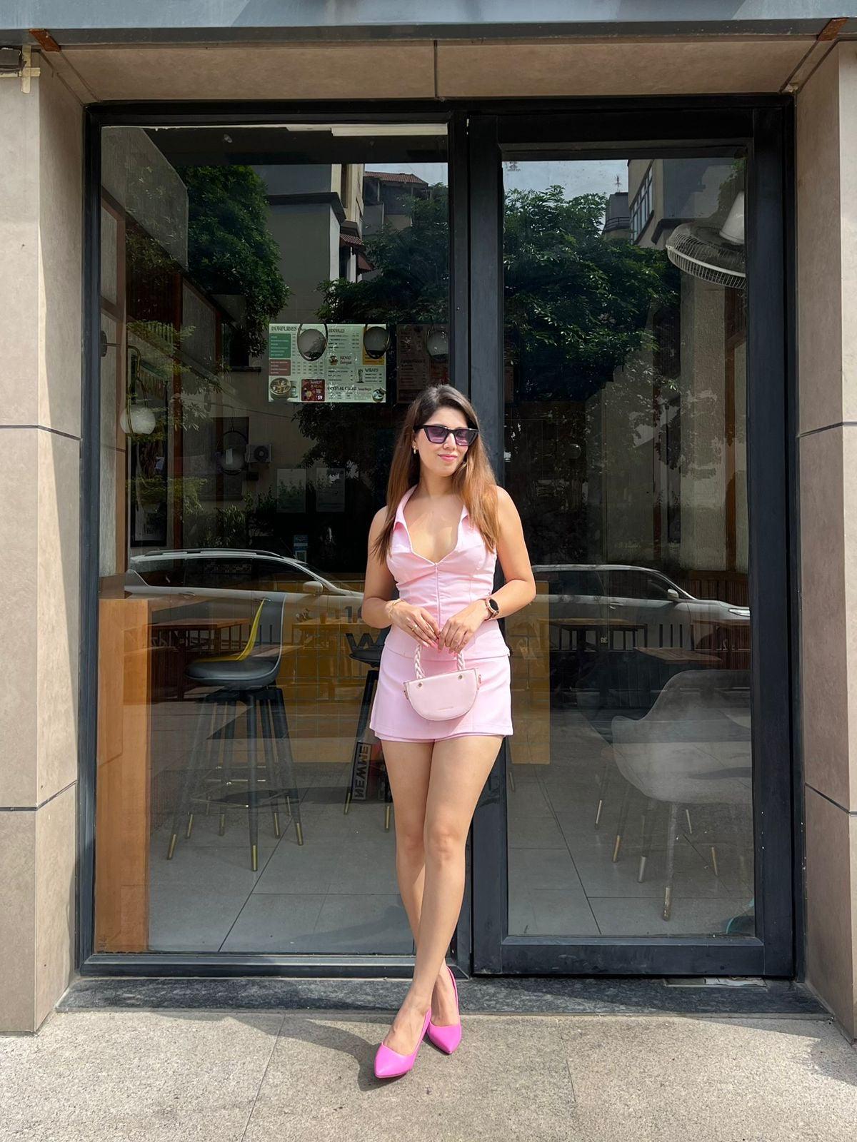 Woman wearing pastel pink halterneck waistcoat and skort set, pink heels, purse, sunglasses, outside a glass storefront.