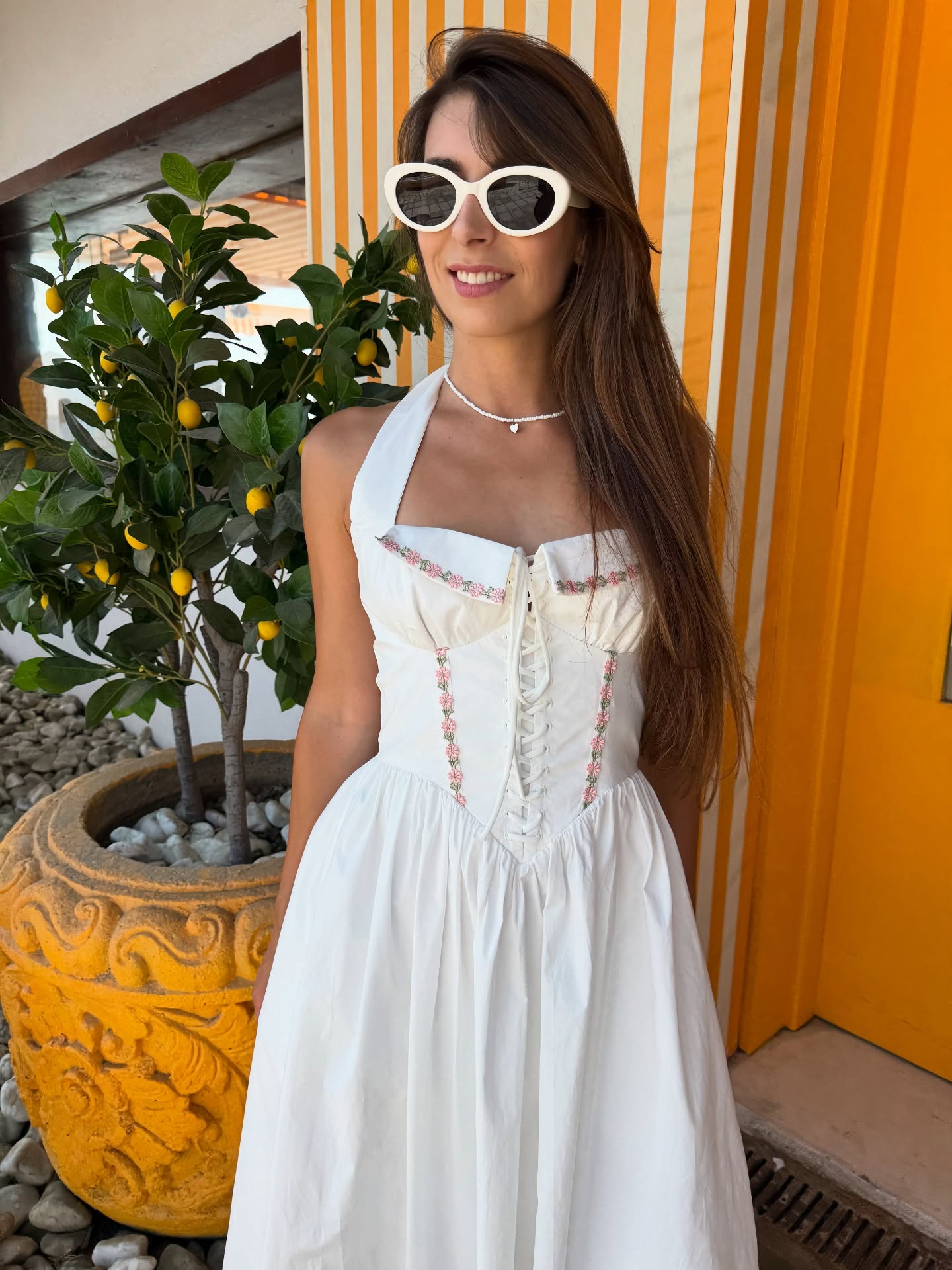 White embroidered corset halter midi dress with pink floral trim and lace-up front, outdoors by yellow striped wall.