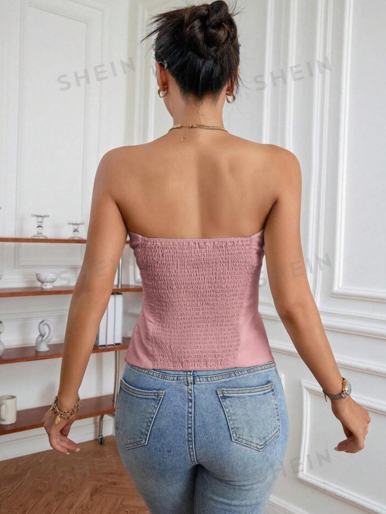 Back view of a woman in a pink smocked strapless bandeau top with light-wash jeans, bun updo, white paneled room.