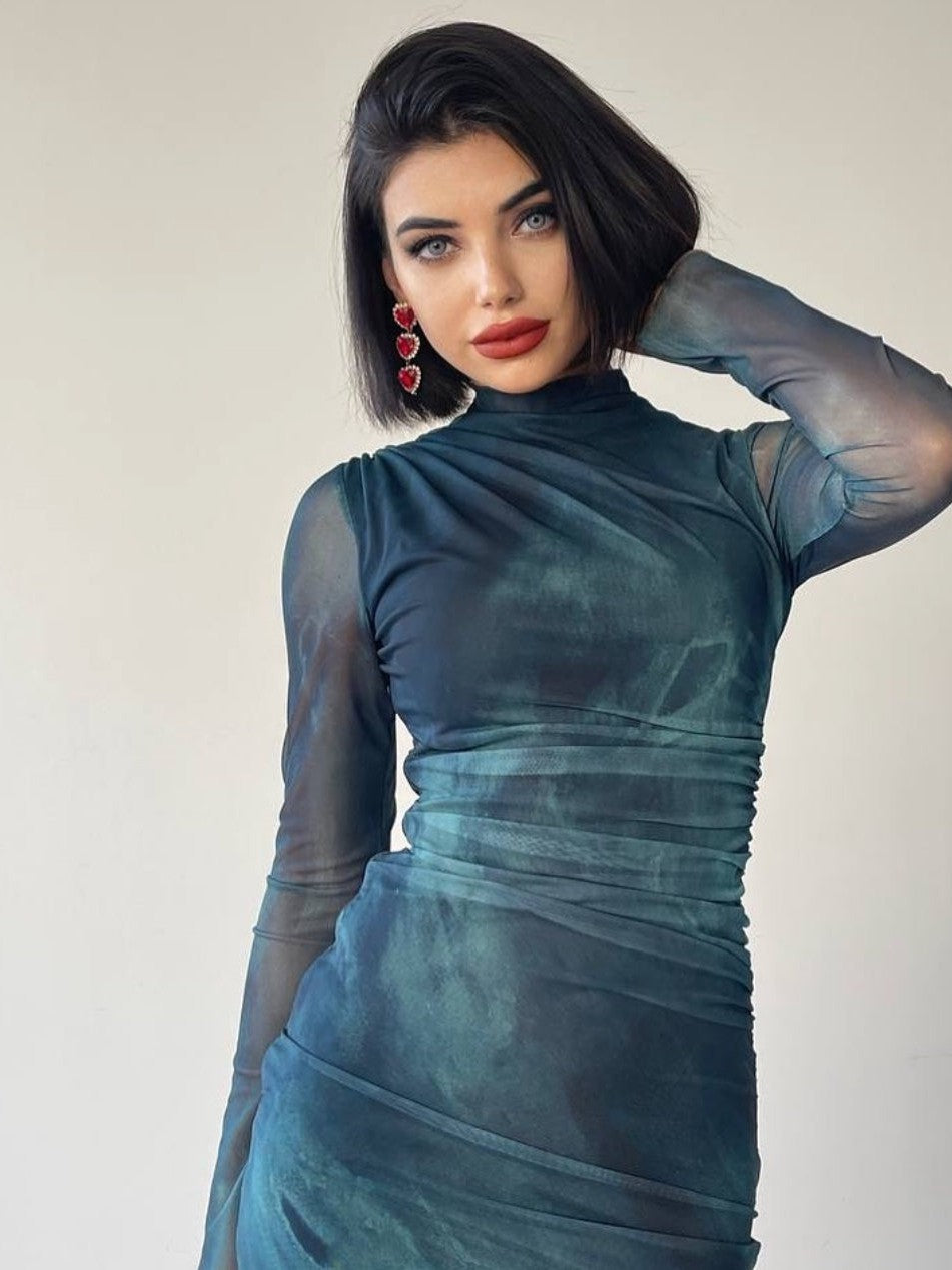 Model wearing teal tie-dye mesh dress with high neck and sheer full sleeves, ruched at the waist; woman has black bob and red earrings.