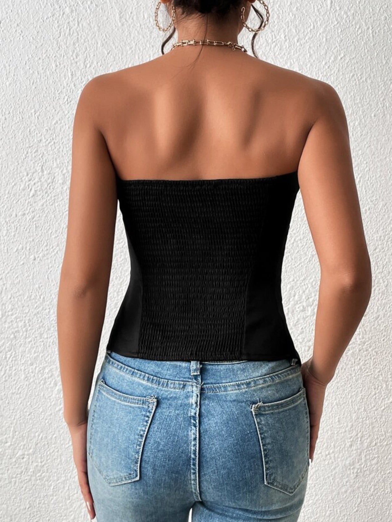 Back view of a woman in a black smocked bandeau top with stretch panels, blue jeans, gold hoop earrings.