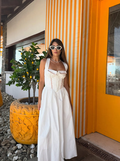 White embroidered corset halter midi dress with pink trim, worn by woman in white sunglasses, against an orange striped wall beside a yellow carved planter.