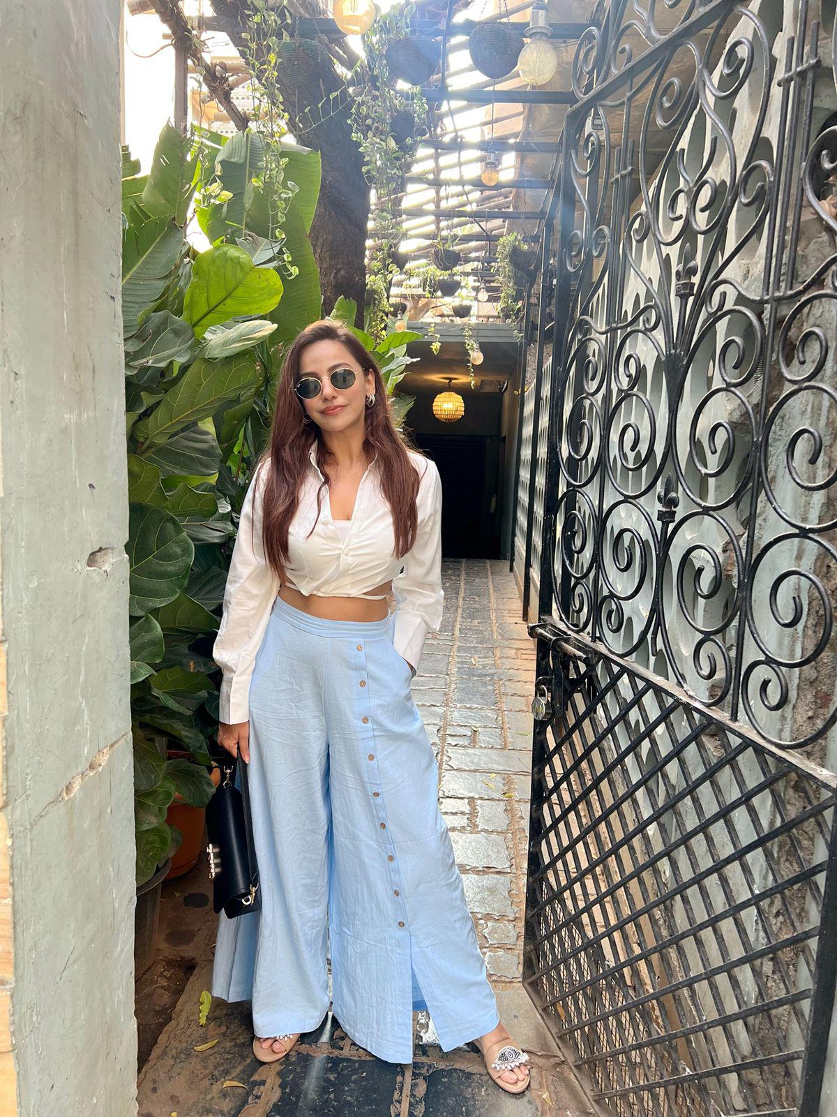 Woman in white crop top with tie-front and light blue linen wide-leg pants, sandals, holding black bag in a leafy alley.