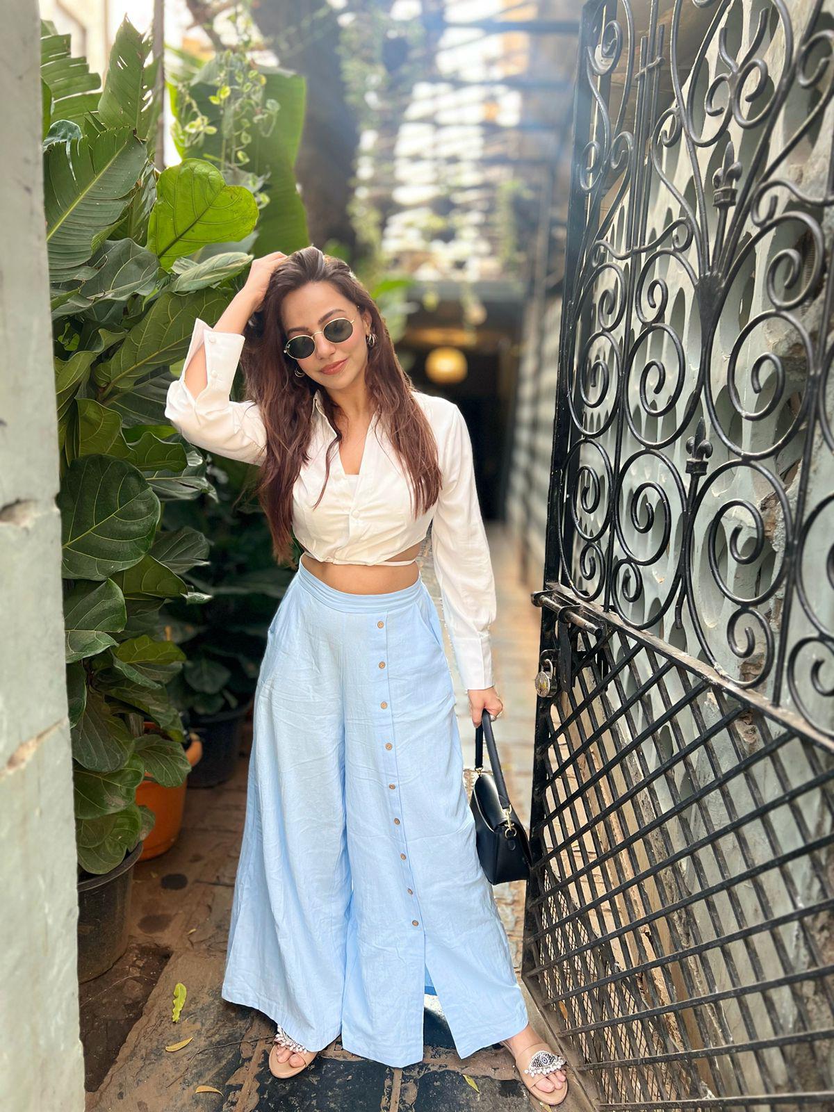 Woman in white crop top and light blue linen button-front wide-leg pants beside ornate gate, wearing sunglasses.