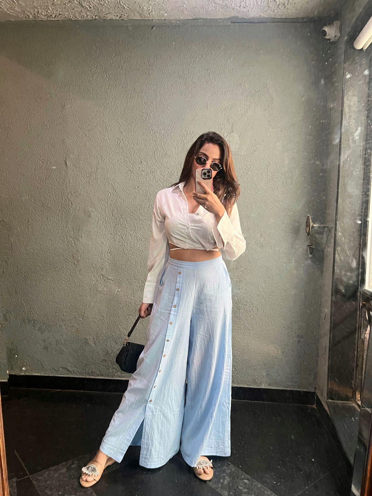 Woman in white cropped blouse and light-blue linen wide-leg button-front pants, holding a small black bag, wearing sunglasses and beige sandals.