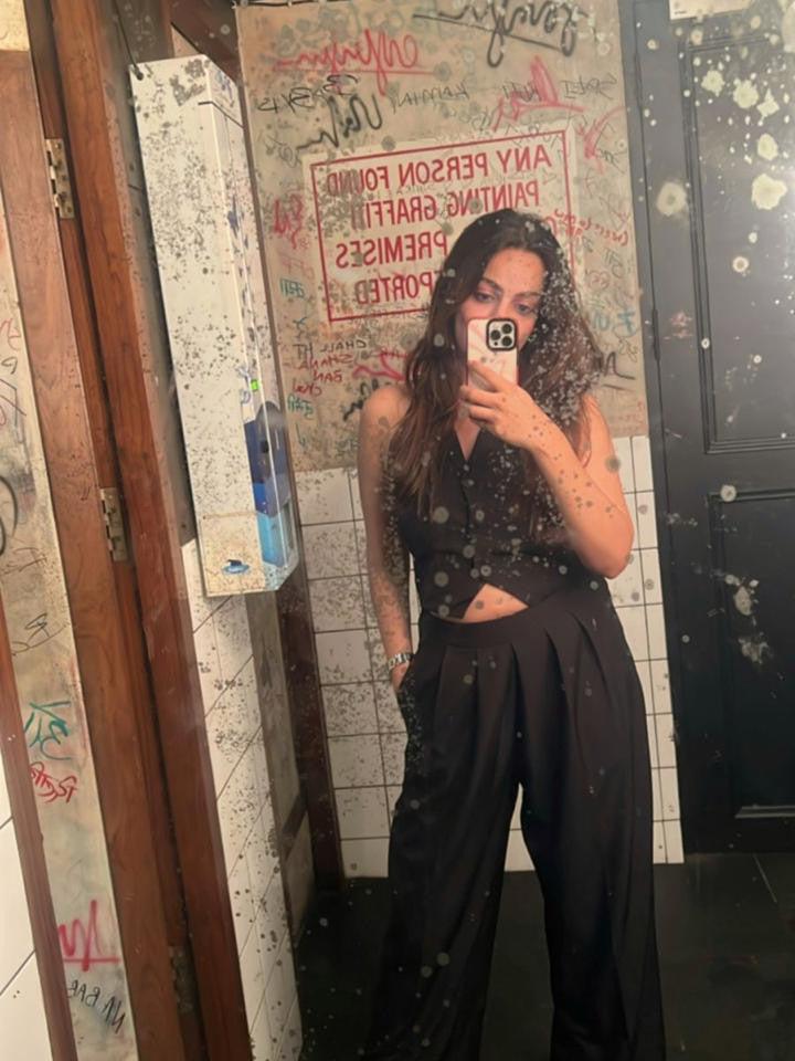 Woman wearing a black cropped waistcoat and matching wide-leg pants, posing in a graffiti-streaked bathroom mirror.