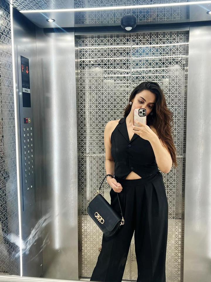 Woman in a black crop-waist coat and wide-leg pants in an elevator, holding a black crossbody bag with gold hardware.