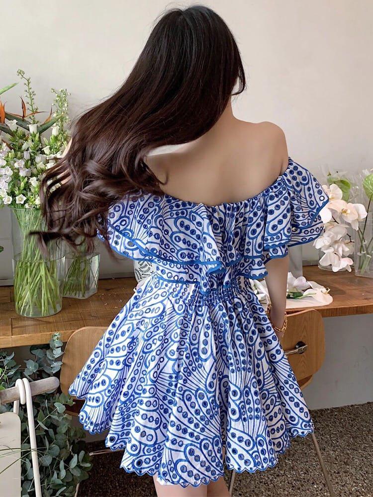 Rear view of a woman in a short off-shoulder blue-and-white embroidered cotton dress with ruffled sleeves and tiered skirt.