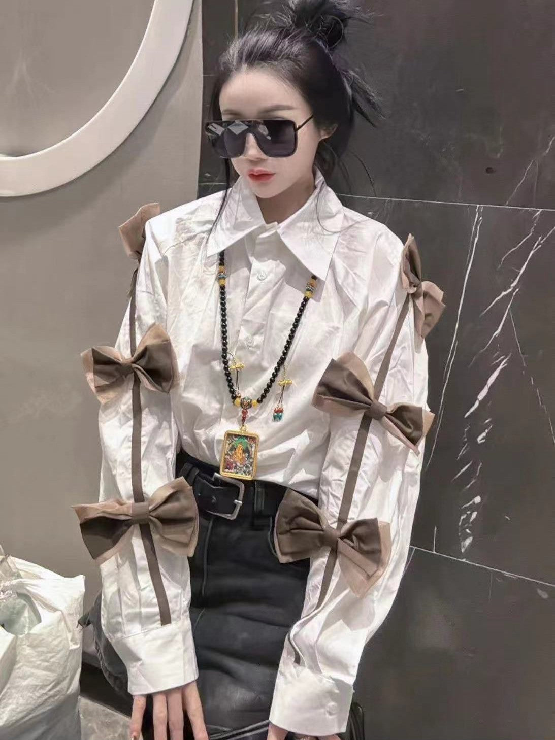 White oversized shirt with large brown bows along the sleeves, paired with a black belt and layered necklace.