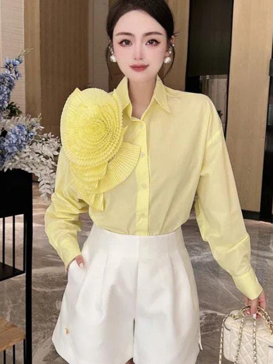 Woman wearing pale yellow shirt with large pleated yellow flower patch on left shoulder, white high-waisted shorts.