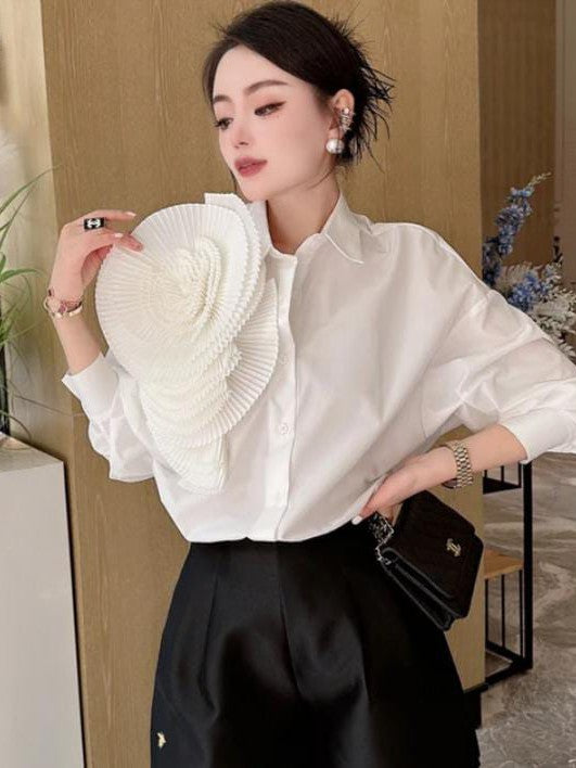 Woman in a white shirt and black high-waisted pants holds a large white pleated circular flower accessory near her face.