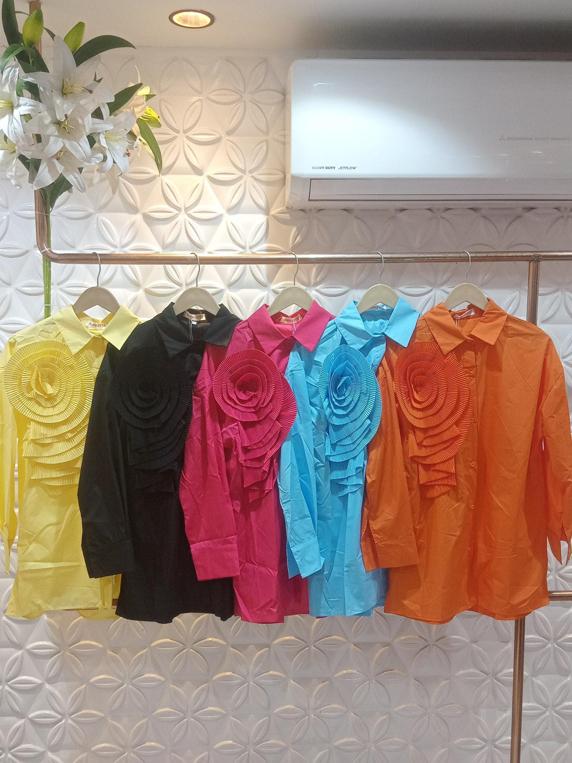 Row of long-sleeve shirts in yellow, black, pink, blue, and orange on a rack, each with a large pleated flower patch.