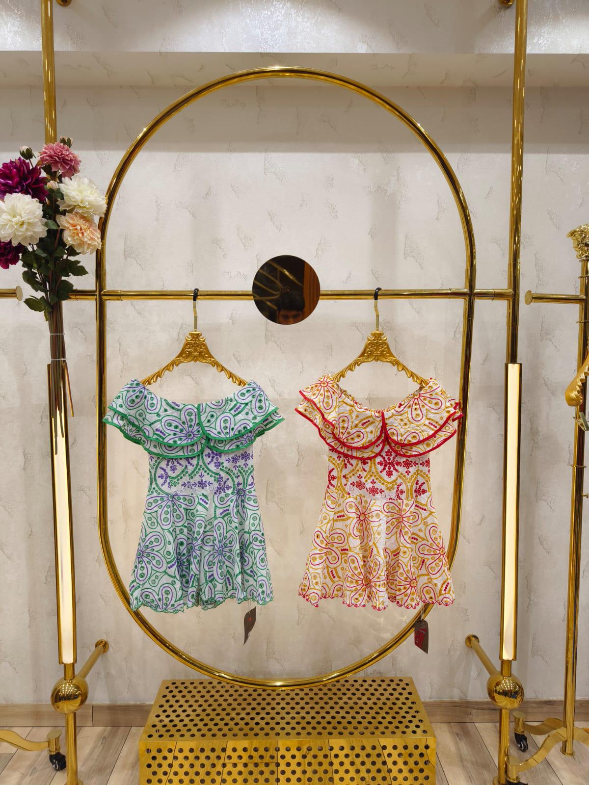 Two off-shoulder embroidered cotton dresses with ruffle necklines, green-on-white and red-on-ivory, on a gold rack.