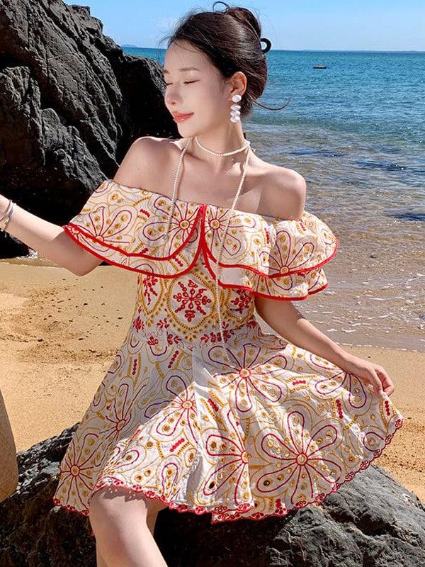 Woman on a rocky beach wearing a short off-shoulder white cotton dress with red-gold embroidered floral pattern.
