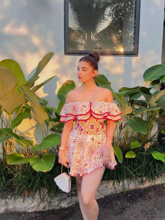 Off-shoulder short embroidered dress with pink floral pattern and red trim, standing by tropical plants.