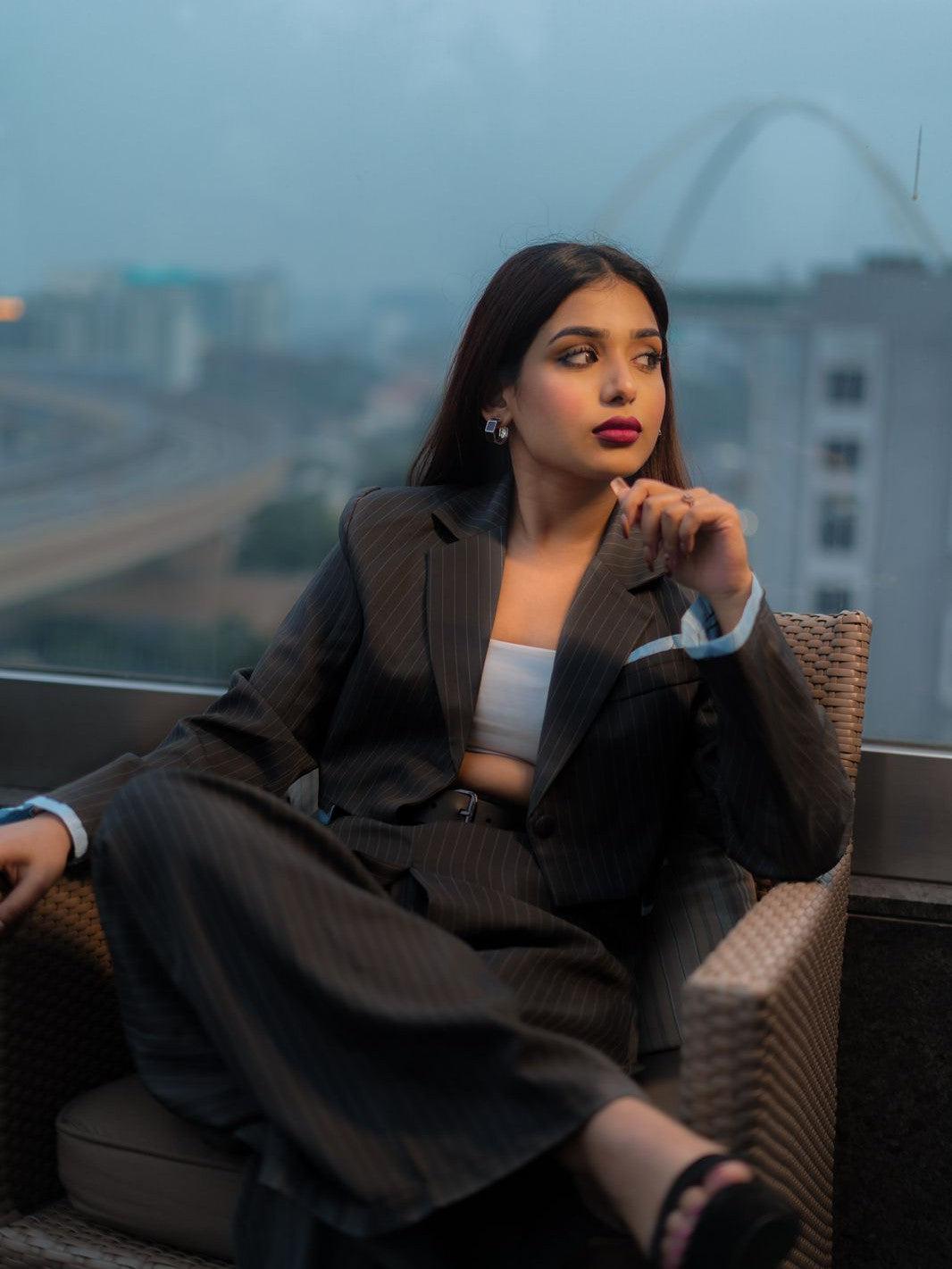 Woman in a dark pinstriped cropped blazer and matching wide-leg pants, white crop top, seated on a wicker chair on a roof.