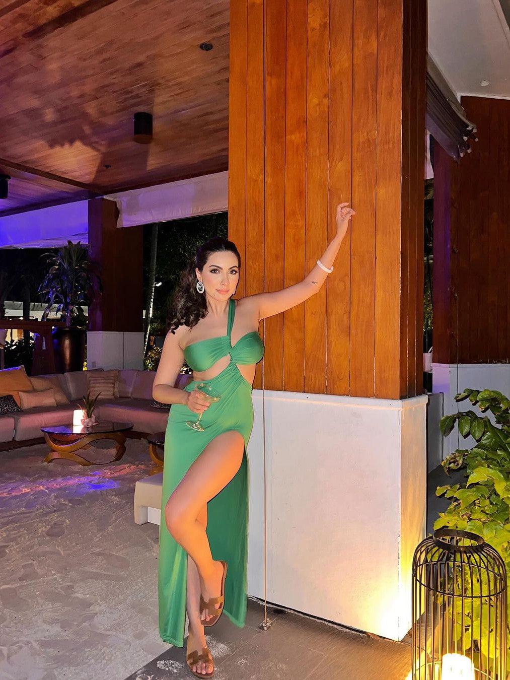 Green bandeau twist, one-shoulder backless maxi with high slit, holding wine glass beside a wooden pillar.