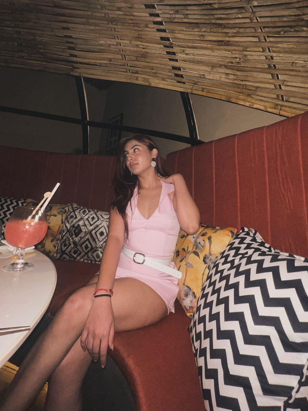Woman wearing a pink halterneck top and matching pink skort with a white belt, seated on a red booth in a stylish lounge.