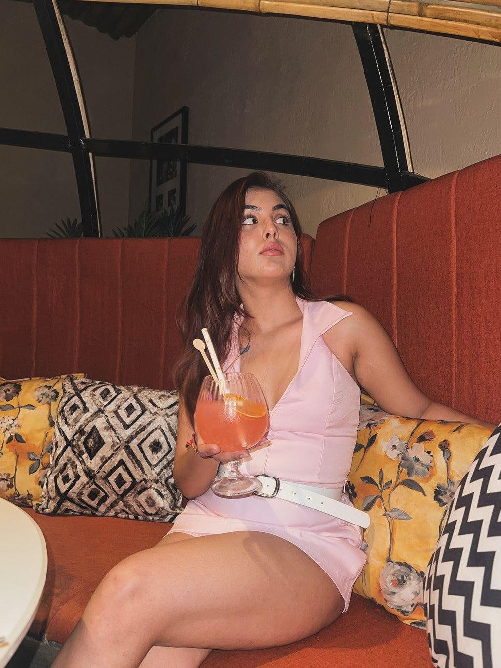 Pink halterneck waist coat with skort set, white belt, worn by a woman on an orange booth, holding a pink cocktail.